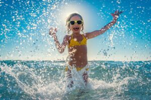 female child splashing in ocean water with yellow sunglasses and bathing suit